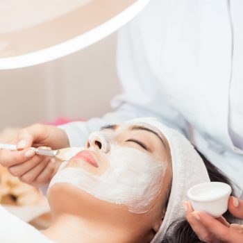Woman receiving a facial treatment, lying under a bright light. Aesthetician applies a white mask with a brush. Relaxed and peaceful atmosphere.